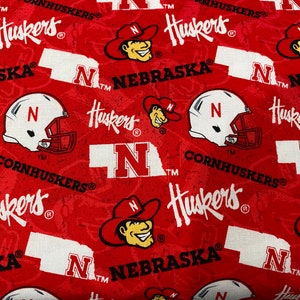 May include: Red fabric with a repeating pattern of the Nebraska Cornhuskers logo, including the team's mascot, a football helmet, and the state of Nebraska outline. The text "Huskers", "Cornhuskers", and "Nebraska" is repeated throughout the fabric.