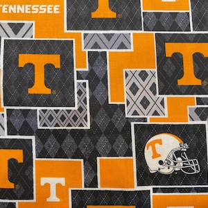 May include: Orange and black fabric with a repeating pattern of the letter "T" and a diamond pattern. The fabric also features a football helmet with the word "Tennessee" on it.