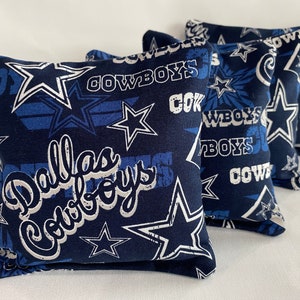 May include: Four blue and white bean bags with the Dallas Cowboys logo and stars. The bean bags are made of fabric and have a soft, plush feel.