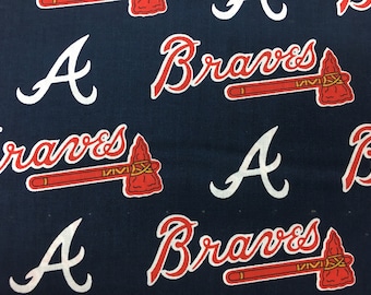 May include: A navy blue fabric with a repeating pattern of the Atlanta Braves logo. The logo features the team's name "Braves" in red script with a tomahawk underneath. The letter "A" in white is also part of the pattern.