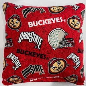 May include: A square throw pillow featuring a red fabric background with a repeating pattern of Ohio State logos, the word BUCKEYES in white text, and illustrations of a football helmet and the Brutus Buckeye mascot character throughout.