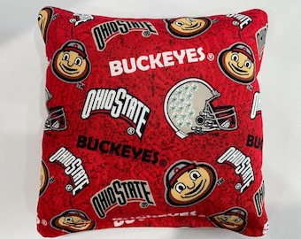 May include: A square throw pillow featuring a red fabric background with a repeating pattern of Ohio State logos, the word BUCKEYES in white text, and illustrations of a football helmet and the Brutus Buckeye mascot character throughout.