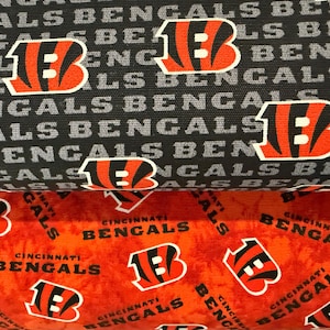 May include: Fabric featuring the Cincinnati Bengals logo in orange, black, and white. The design includes the team's logo and the word "BENGALS" repeated across a black and orange background.