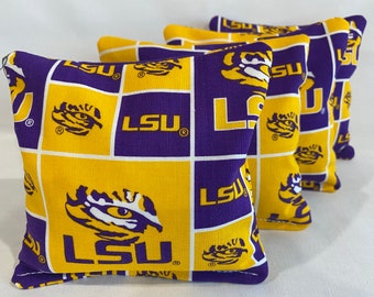 May include: Four purple and gold bean bags with the LSU logo and tiger paw print. The bean bags are made of fabric and have a white border.