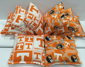 Op de afbeelding: Een verzameling oranje en witte zitzakken. Sommige tassen hebben de letter "T" in een rasterpatroon, terwijl andere logo's en tekst van "Tennessee Volunteers" weergeven. De zitzakken zijn waarschijnlijk voor een spel of decoratief gebruik.
