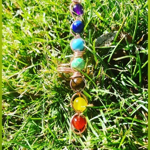 May include: A copper wire wrapped ring with seven colorful gemstones. The gemstones are purple, blue, turquoise, green, brown, yellow, and red. The ring is lying on a bed of green grass.