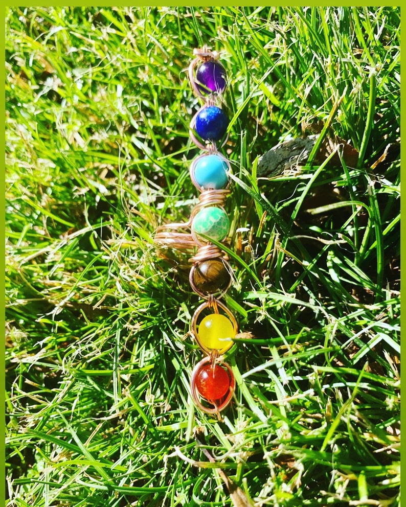 May include: A copper wire wrapped ring with seven colorful gemstones. The gemstones are purple, blue, turquoise, green, brown, yellow, and red. The ring is lying on a bed of green grass.