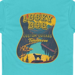 May include: A turquoise blue t-shirt with a guitar-shaped graphic on the back. The graphic features a sunset scene with mountains, a lake, and a black dog. The text "Lucky Dog Custom Guitars Tennessee" is printed on the guitar.