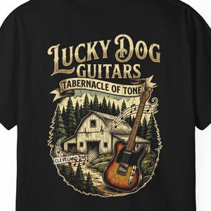 Op de afbeelding: Zwart t-shirt met een afbeelding van een gitaar, schuur en bomen. De tekst luidt "Lucky Dog Guitars Tabernacle of Tone Cleveland, Ohio". De gitaar heeft een bruine body en een zwarte hals.