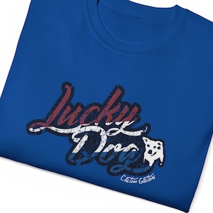 May include: A royal blue t-shirt with a distressed graphic design that says "Lucky Dog" in red, white, and blue with a white silhouette of a dog's head. The text "Custom Guitars" is printed in white below the graphic.