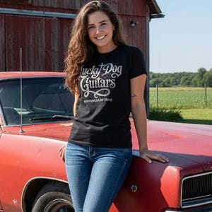 Puede incluir: Un coche rojo clásico vintage con una parrilla negra y detalles cromados. Una mujer con una camiseta negra con el texto "Lucky Dog Guitars" y vaqueros azules está de pie junto al coche. El fondo incluye un granero rojo y campos verdes.