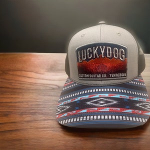 May include: A gray and black trucker hat with a black mesh back. The front of the hat features a gray patch with the text "LUCKYDOG CUSTOM GUITAR CO. TENNESSEE" and a red, orange, and yellow mountain scene.