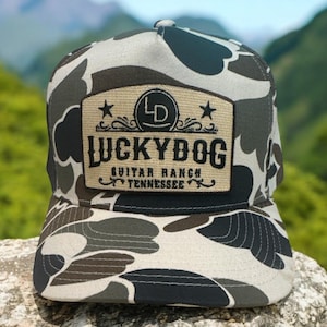 Peut inclure: Casquette de baseball camouflage avec un écusson portant l'inscription "LUCKYDOG GUITAR RANCH TENNESSEE". La casquette a une visière incurvée et une couronne structurée. L'arrière-plan montre un paysage de montagne flou.