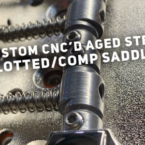 Op de afbeelding: Close-up van een gitaarbrug met aangepaste CNC-gefreesde, verouderde stalen gesleufde/comp-zadels. De zadels zijn zilverkleurig en hebben een textuur.