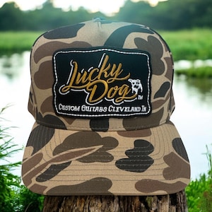 Peut inclure: Casquette de baseball camouflage avec une visière plate. Le chapeau comporte un écusson noir avec les mots "Lucky Dog" en lettres dorées et la phrase "Custom Guitars Cleveland IN". La casquette repose sur une souche d'arbre.