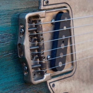 Op de afbeelding: Close-up van een zilveren gitaarbrug met zes snaren en een zwarte pickup. De brug heeft een getextureerd oppervlak en is gemonteerd op een lichtblauwe en witte gitaarbody.