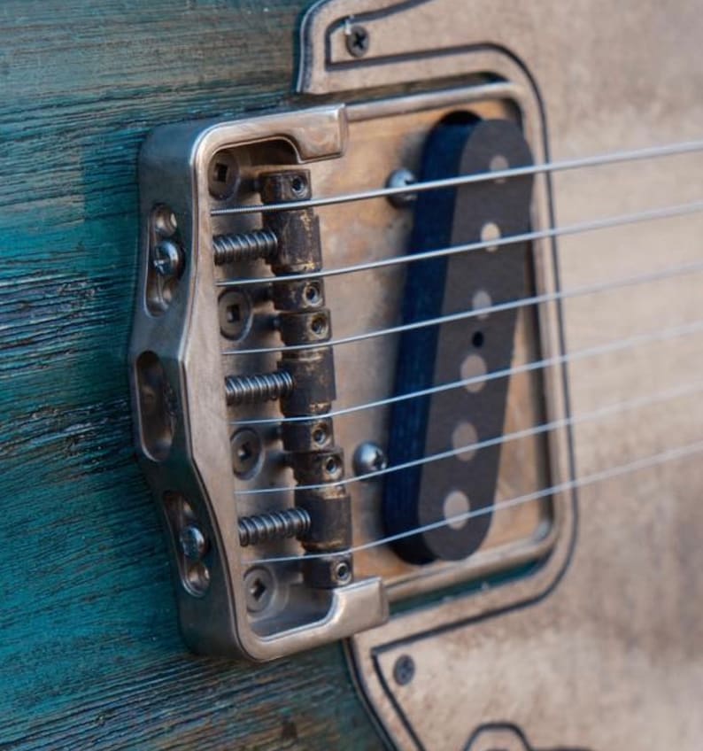 Op de afbeelding: Close-up van een zilveren gitaarbrug met zes snaren en een zwarte pickup. De brug heeft een getextureerd oppervlak en is gemonteerd op een lichtblauwe en witte gitaarbody.