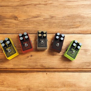 May include: Five guitar effects pedals on a wooden surface. The pedals are all different colors and have the words "Beef Cake" printed on them.
