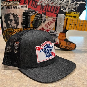 May include: A black denim trucker hat with a mesh back and a curved brim. The hat features a patch with the text "LUCKY DOG CUSTOM GUITARS" in a red, white, and blue design. A guitar with stickers is in the background.
