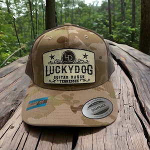 May include: A brown and tan camouflage trucker hat with a brown leather patch that says "Lucky Dog Guitar Ranch Tennessee" with a star on each side of the patch. The hat has a black mesh back and a black snap closure.