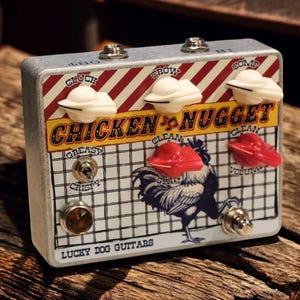 May include: A silver guitar effects pedal with a red and white striped top. The pedal features white knobs shaped like UFOs and red knobs. The text "Chicken Nugget" is displayed prominently, along with a rooster illustration. The brand name "Lucky Dog Guitars" is also visible.