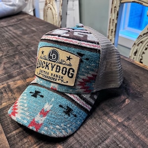 May include: A trucker hat with a turquoise, red, and white patterned design on the front panels and bill. The hat features a mesh back and a patch that reads "Lucky Dog Guitar Ranch Tennessee."