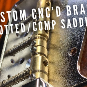 Op de afbeelding: Close-up van een gitaarbrug met aangepaste CNC-gefreesde messing gesleufde/comp-zadels. De zadels zijn goudkleurig en hebben een textuur. De brug is gemaakt van verchroomd metaal.