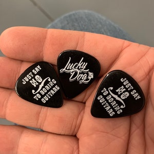 May include: Three black guitar picks. One pick has the text "Just Say No To Boring Guitars" and another pick has the text "Lucky Dog". The third pick has the text "Just Say No To Boring Guitars".