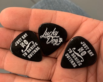 May include: Three black guitar picks. One pick has the text "Just Say No To Boring Guitars" and another pick has the text "Lucky Dog". The third pick has the text "Just Say No To Boring Guitars".