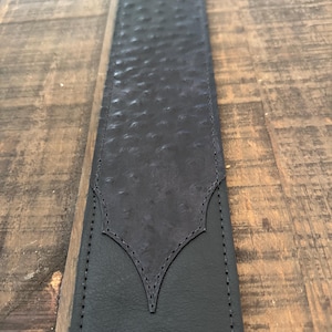 May include: Black leather guitar strap with a textured, embossed pattern. The strap has a decorative, pointed design at the top.