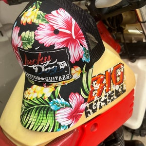 Puede incluir: Gorra de camionero negra con un estampado floral de flores rojas, rosas, amarillas y azules y hojas verdes. La gorra tiene una parte trasera de malla negra y un parche que dice "Lucky Dog Custom Guitars".