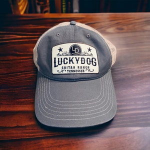 May include: A gray and beige mesh baseball cap with a white patch that says "Lucky Dog Guitar Ranch Tennessee" with a stylized "LD" logo inside a circle.
