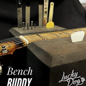 May include: A close-up of a guitar with a black body and a wooden neck. A black foam block holds various guitar tools, including metal files and a yellow pick. The text "Bench Buddy" and "Lucky Dog" are visible.