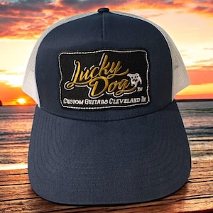 May include: A navy blue and white trucker hat with a black patch that says "Lucky Dog Custom Guitars Cleveland, OH" in gold lettering. The patch also features a small white dog silhouette.