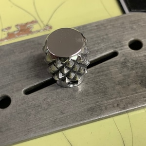 Puede incluir: Un pomo de color plateado en forma de diamante con una superficie lisa y reflectante. El pomo est&aacute; montado en una placa de metal.
