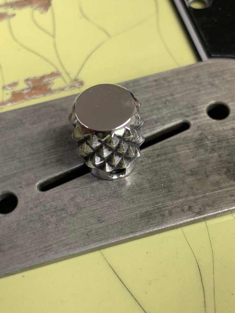 Puede incluir: Un pomo de color plateado en forma de diamante con una superficie lisa y reflectante. El pomo est&aacute; montado en una placa de metal.
