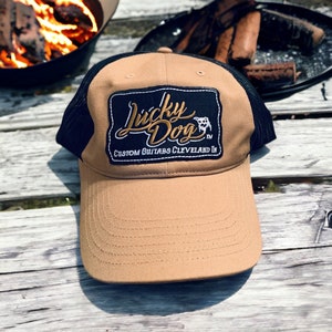 May include: A tan and black trucker hat with a black patch that says "Lucky Dog" in gold lettering. The patch also says "Custom Guitars Cleveland, OH" in white lettering.