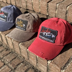 May include: Three distressed baseball caps in different colors: blue, gray, and red. Each cap features a black patch with the text "Lucky Dog Custom Guitars" in white and yellow lettering.