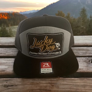 Op de afbeelding: Een zwarte en grijze truckercap met een grijs voorpaneel en een zwart mesh achterpaneel. Het voorpaneel heeft een zwart en gouden geborduurd patch met de tekst "Lucky Dog Custom Guitars Cleveland, OH" en een klein hondensilhouet. De pet heeft ook een zwart-wit label met de tekst "Richardson Flat Bill Snapback" aan de voorkant.