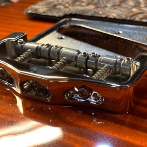 Może przedstawiać: Chromowany mostek gitarowy z regulowanymi siodełkami i blokadą strun. Mostek jest przeznaczony do gitary sześciostrunowej i ma klasyczny wygląd.