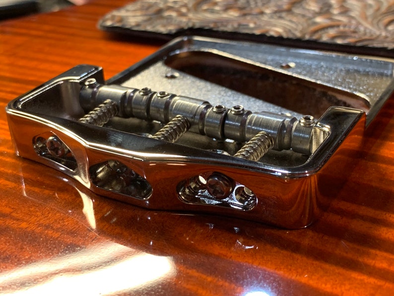 Op de afbeelding: Een verchroomde gitaarbrug met verstelbare zadels en een snaarhouder. De brug is ontworpen voor een gitaar met zes snaren en heeft een klassieke uitstraling.