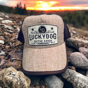 May include: A brown and beige trucker hat with a black mesh back. The hat has a patch on the front that says "LUCKYDOG GUITAR RANCH TENNESSEE" with a star on each side of the patch.