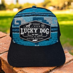 May include: A black and blue trucker hat with a patterned top and a mesh back. The hat features a patch with the text "LUCKY DOG EST 2015 CUSTOM GUITARS" and a dog silhouette. The hat is sitting on a brick surface.