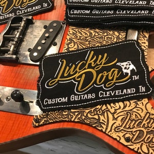 May include: Close-up of a guitar with a black and gold "Lucky Dog" patch. The patch has the text "Custom Guitars Cleveland IN" on it. The guitar is a reddish-brown color with a floral pattern on the body.