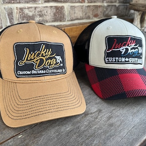 Può includere: Due cappellini da camionista con retro in rete nera. Il cappellino a sinistra è marrone con una toppa nera che dice "Lucky Dog Custom Guitars Cleveland, OH". Il cappellino a destra è bianco con una visiera a scacchi neri e rossi e una toppa nera che dice "Lucky Dog Custom Guitars".