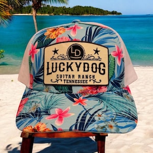 Puede incluir: Una gorra de camionero azul claro y blanca con estampado floral tropical. La gorra tiene un parche marrón con el texto "Lucky Dog Guitar Ranch Tennessee" y una estrella a cada lado del parche.