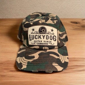 May include: A camouflage baseball cap with a brown leather patch that says "Lucky Dog Guitar Ranch Tennessee" with a star on each side of the patch.