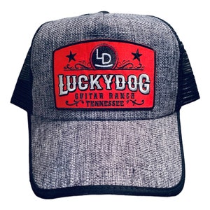 May include: A gray and black trucker hat with a red patch that says "Lucky Dog Guitar Ranch Tennessee" with a star on each side of the patch.
