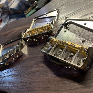 Op de afbeelding: Drie verchroomde gitaarbrugplaten met goudkleurige zadels en schroeven. De platen liggen op een donker houten oppervlak.