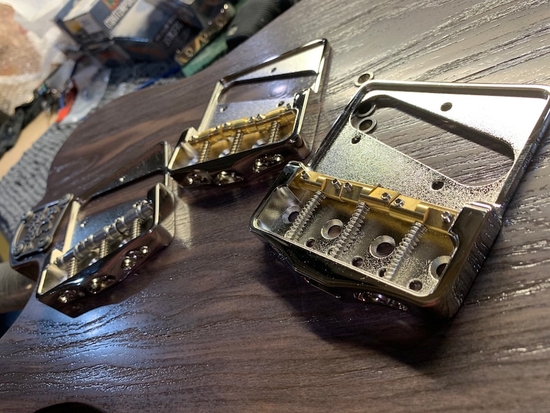 Op de afbeelding: Drie verchroomde gitaarbrugplaten met goudkleurige zadels en schroeven. De platen liggen op een donker houten oppervlak.